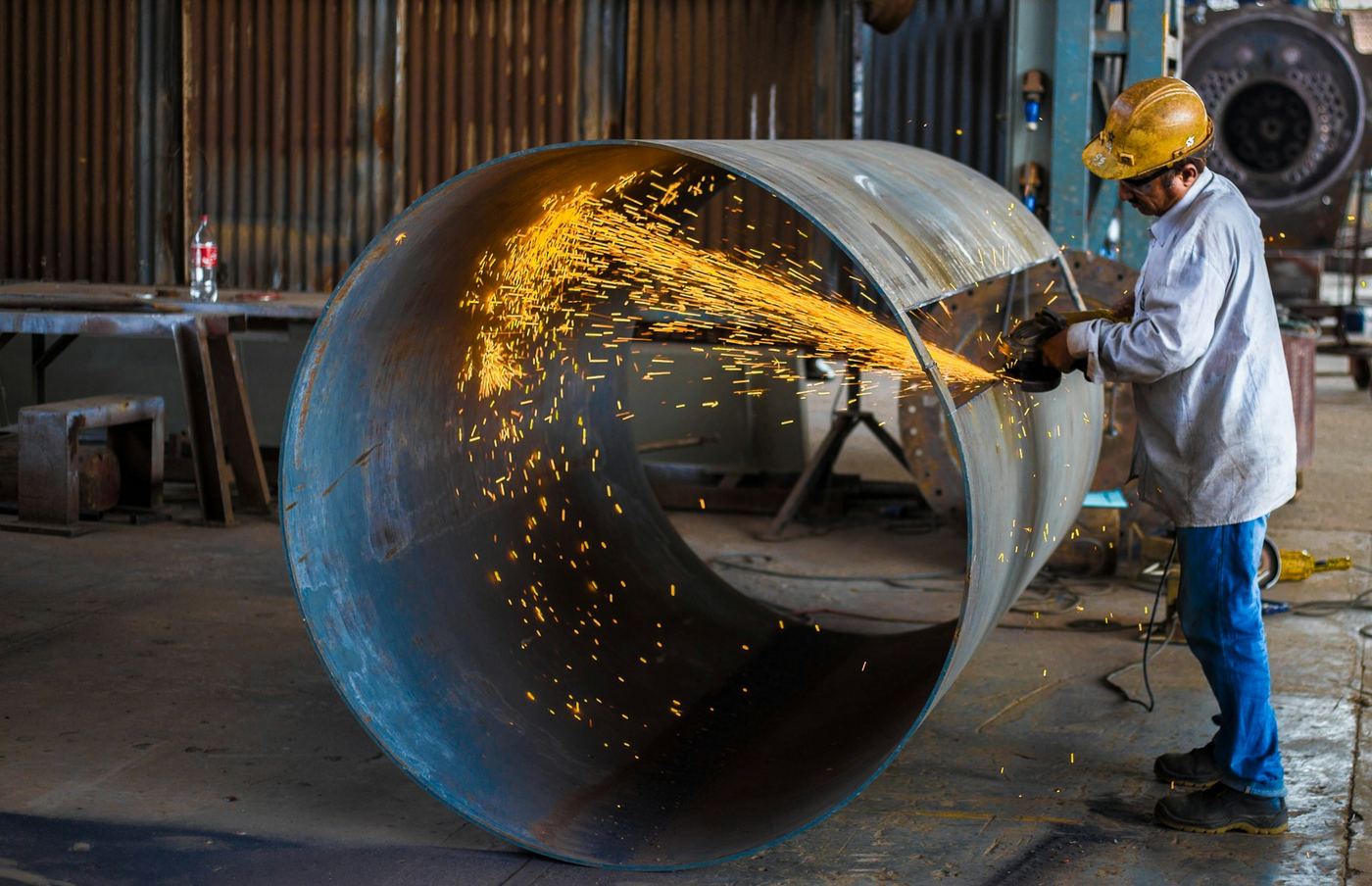 Fabricator in a hard hat grinding a large steel pipe section in a workshop